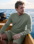Man wearing a green long-sleeve shirt and shorts sitting on a boat with water in the background.