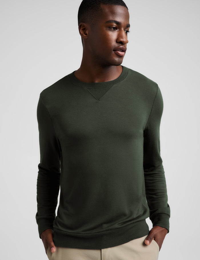 A man wearing a dark olive green long-sleeve crewneck sweater, paired with beige trousers, photographed against a neutral light gray background.