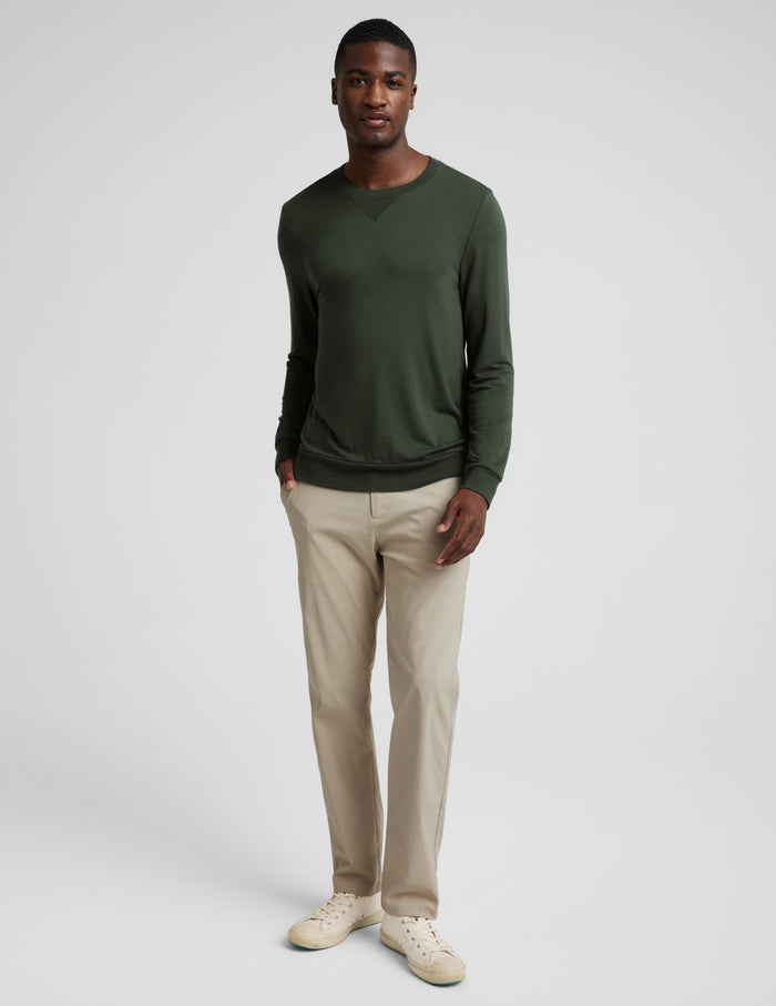 A man wearing a dark olive green long-sleeve crewneck sweater, paired with beige trousers, photographed against a neutral light gray background.