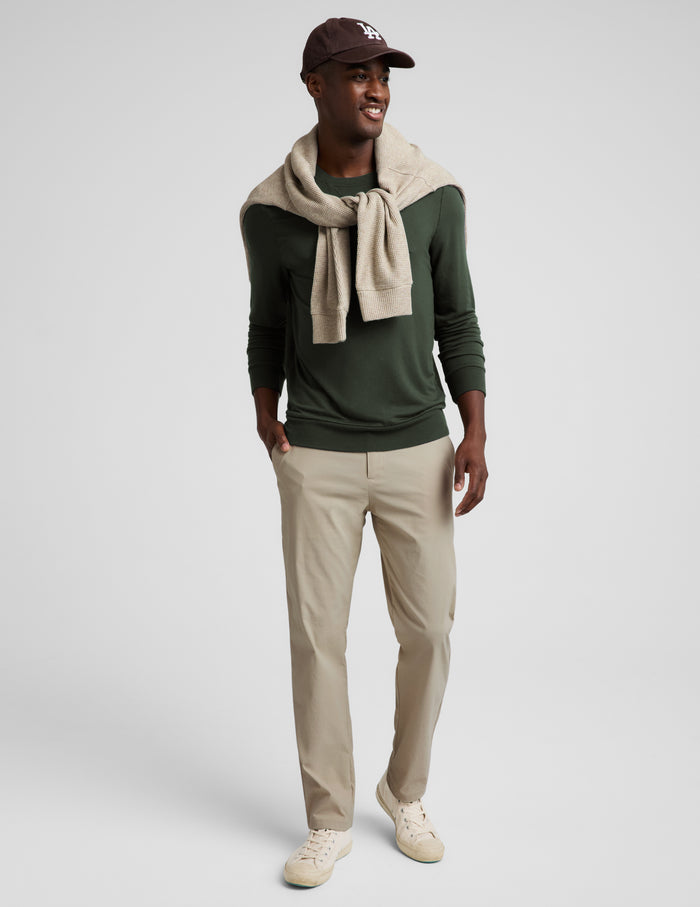 A man modeling a dark green crewneck sweater, beige trousers, a beige knit scarf, and a dark brown baseball cap against a light gray studio background.