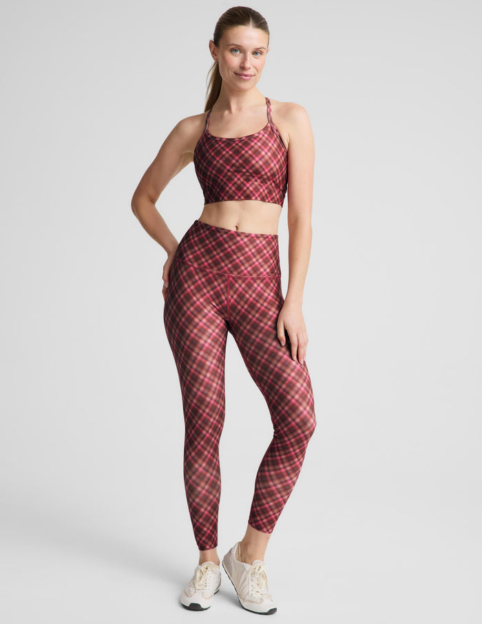 A woman modeling a matching athletic set featuring a red and pink plaid sports bra and high-waisted leggings against a light gray studio background.