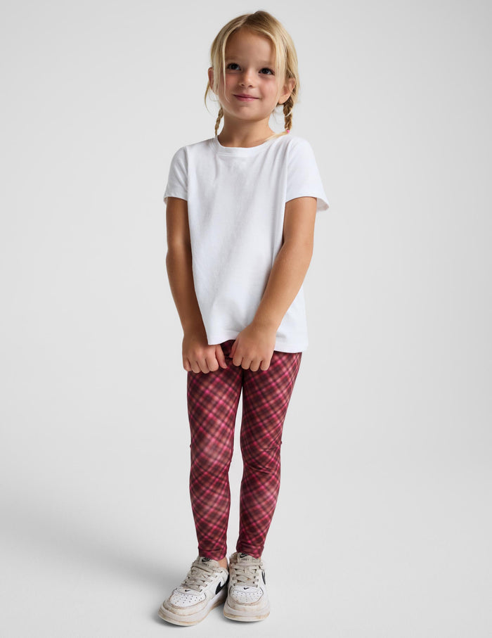 A young girl with blonde braids stands against a light gray studio background, wearing a plain white short-sleeve t-shirt, red and pink plaid leggings, and white sneakers.