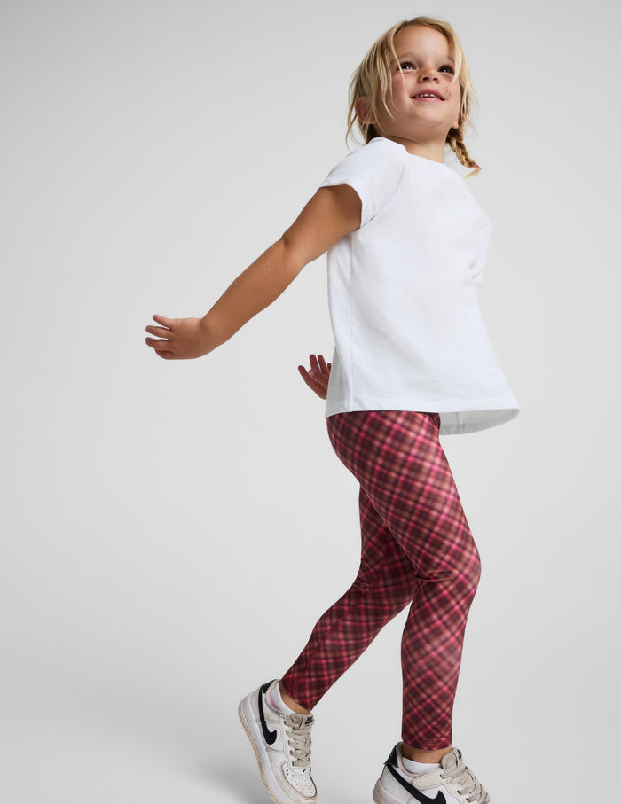 A young girl wearing red and black plaid leggings, a white t-shirt, and white sneakers, captured in a dynamic pose against a light gray studio background.