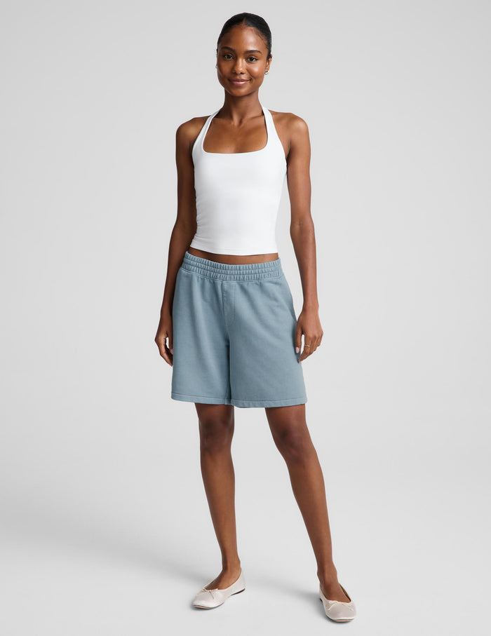A woman modeling a white halter tank top and light blue bermuda shorts against a light gray studio background.