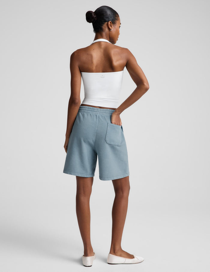 A woman modeling a white halter tank top and light blue bermuda shorts against a light gray studio background.