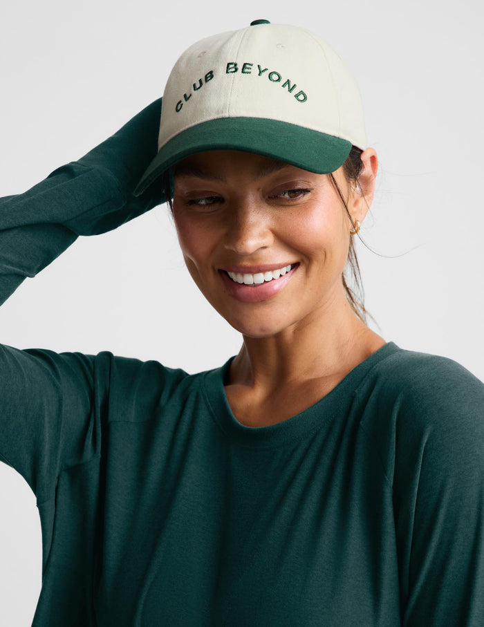A woman wearing a dark green long-sleeve top and a cream and dark green baseball cap with the text 'CLUB BEYOND' embroidered on the front, posed against a light gray background.