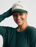 A woman wearing a dark green long-sleeve top and a cream and dark green baseball cap with the text 'CLUB BEYOND' embroidered on the front, posed against a light gray background.