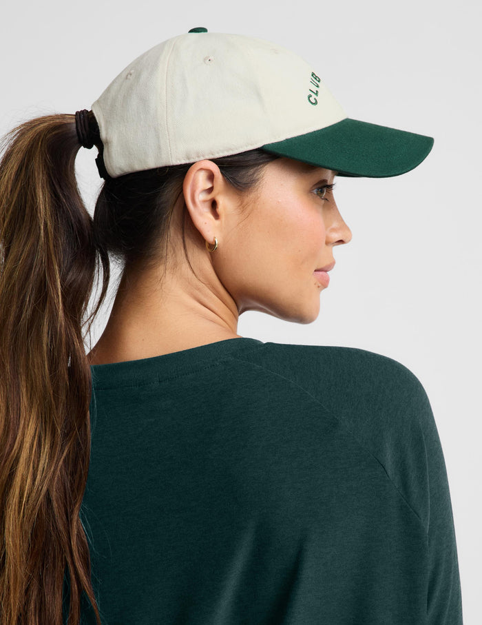A woman shown from the back and side wearing a baseball cap with an off-white crown and a dark green brim. The cap has green embroidered text reading 'CLUB' on the side.