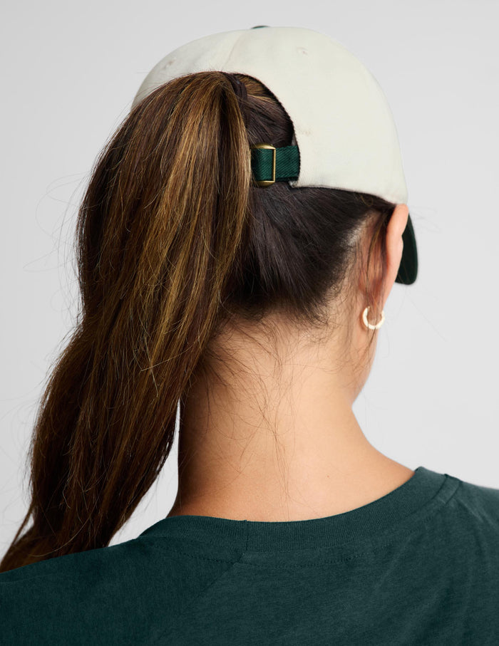 Rear view of a woman wearing a light beige baseball cap with a dark green adjustable strap and gold buckle, paired with a dark green t-shirt against a light gray background.