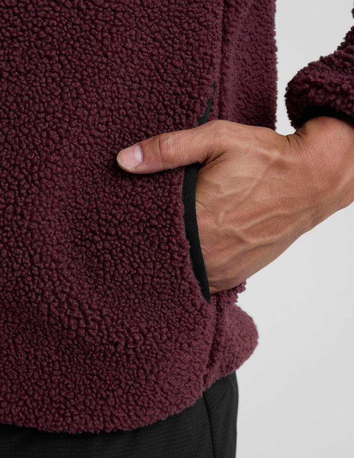 model wearing a burgundy fleece men's zip-up jacket on a gray background.