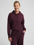A woman modeling a matching ribbed lounge set in deep burgundy, featuring a midway length long-sleeve hoodie with drawstrings and coordinating drawstring pants, against a solid light gray background.