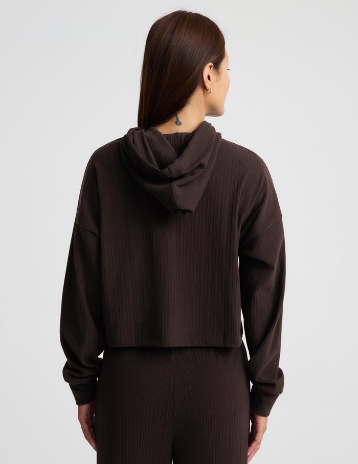 A woman modeling a dark brown ribbed knit hoodie and matching pants against a light gray studio background.