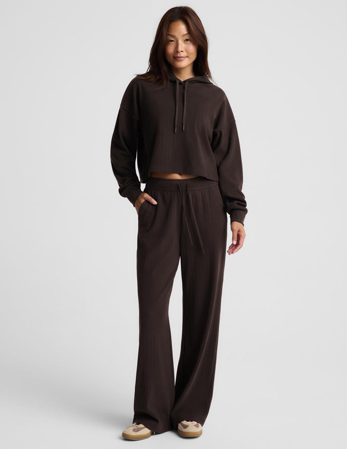 A woman modeling a matching dark brown lounge set consisting of a cropped long-sleeve hoodie with drawstrings and matching wide-leg sweatpants, photographed against a light gray studio background.