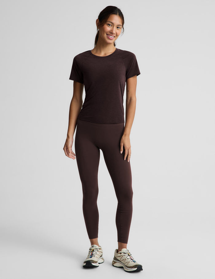 A woman modeling a dark brown short-sleeve athletic t-shirt and matching dark brown high-waisted leggings, paired with beige and black running shoes, against a light gray studio background.
