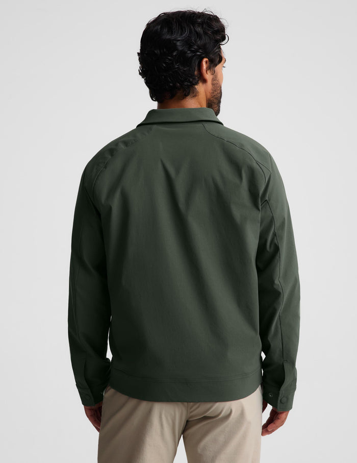 A man viewed from behind wearing an olive green long-sleeve jacket and beige trousers, photographed against a plain light gray studio background.