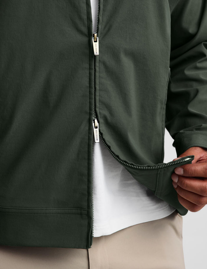 Close-up of a person wearing a dark olive green zip-front jacket over a white shirt and beige pants, with the hand lifting the hem of the jacket to show the zipper.