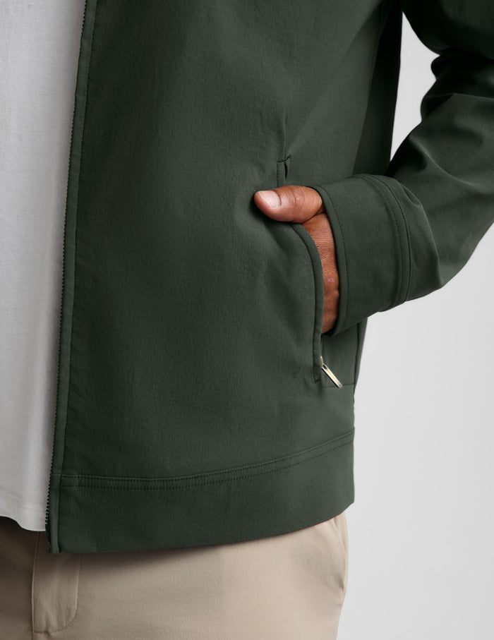 Close-up of a person wearing a dark olive green zip-front jacket over a white shirt and beige pants, with the hand tucked into a side pocket.