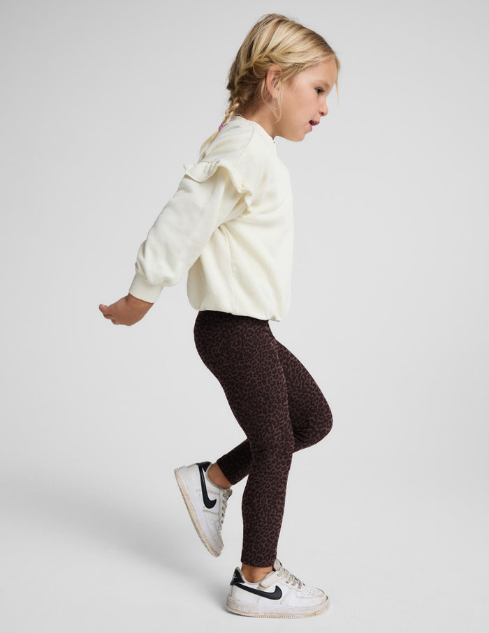 A young girl posing in dark brown leopard-print leggings, a cream puff-sleeve sweatshirt, and white sneakers against a light gray studio background.