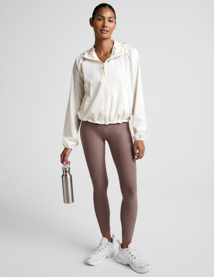Woman wearing a white jacket and brown leggings, holding a water bottle on a light gray background.