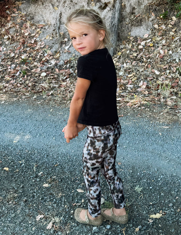 A young girl posing outdoors on a gravel path, wearing a black short-sleeve t-shirt and leopard-print leggings.