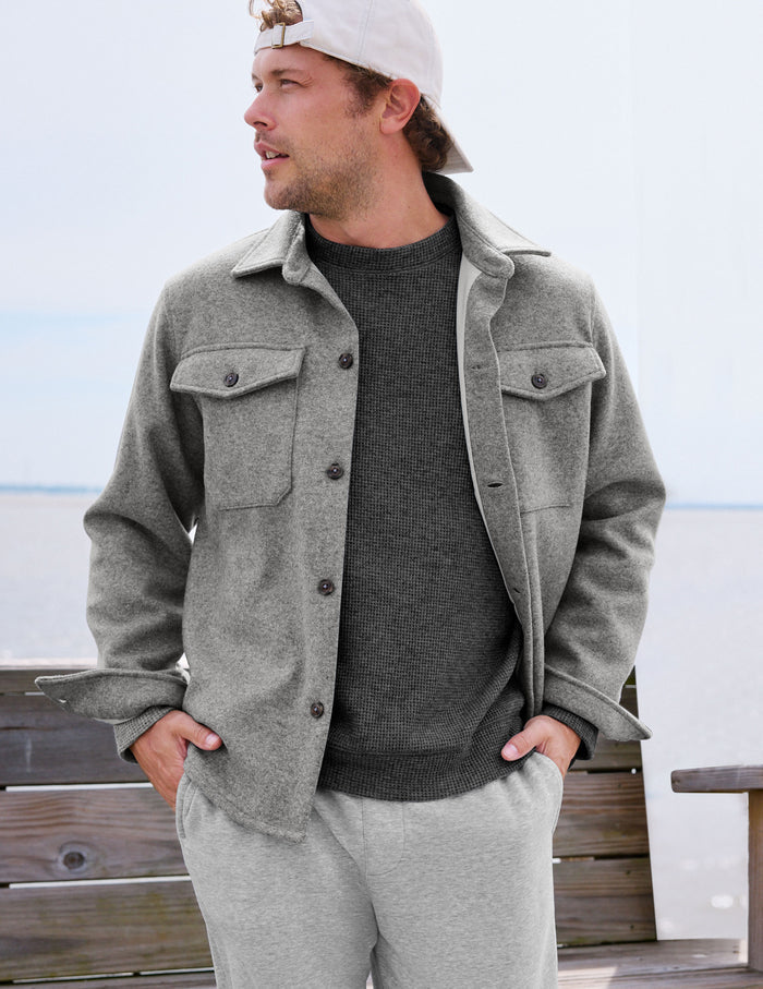 A man wearing a light gray wool-blend button-up overshirt with chest flap pockets, a dark gray knit sweater, and light gray sweatpants, standing on a wooden boardwalk with water in the background.