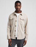 A man wearing a light beige textured button-up overshirt over a white t-shirt and dark gray trousers, photographed against a neutral light gray background.