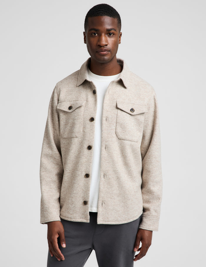 A man wearing a light beige textured button-up overshirt over a white t-shirt and dark gray trousers, photographed against a neutral light gray background.