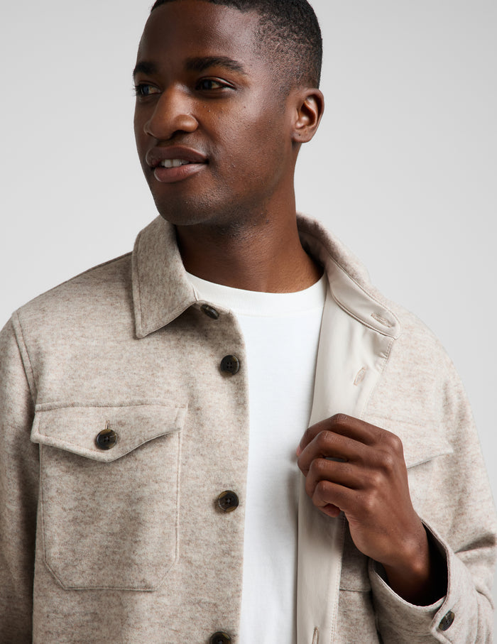 A man wearing a light beige textured overshirt jacket over a white t-shirt, photographed against a neutral light gray background.