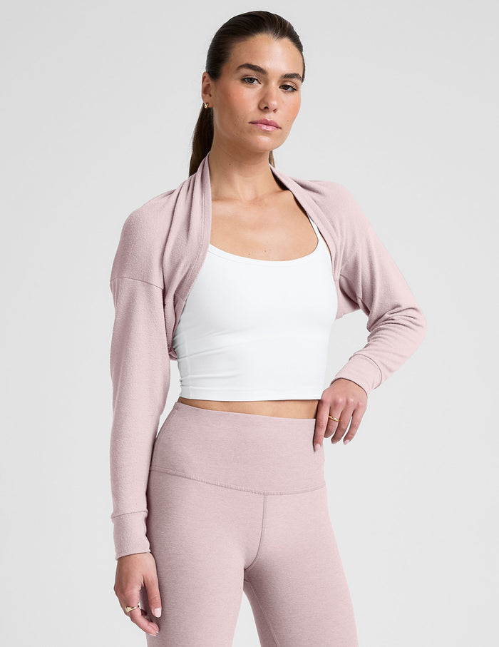 woman wearing a pink long-sleeve crop shrug and leggings on a plain background.