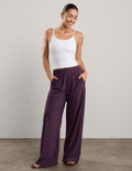 A woman shown from the back wearing a white tank top and dark purple wide-leg pants, standing against a neutral studio backdrop.