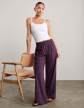 A woman modeling a white camisole top and high-waisted, wide-leg purple pants, standing next to a mid-century modern wooden chair against a neutral studio background.