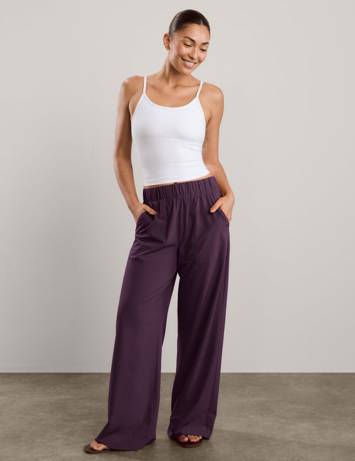 A woman shown from the back wearing a white tank top and dark purple wide-leg pants, standing against a neutral studio backdrop.