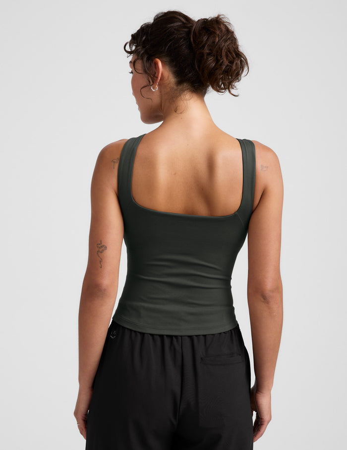 Woman wearing a green sleeveless top and black pants on a light gray background.