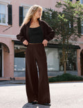 A woman stands outdoors on a street wearing a dark brown bomber jacket, matching wide-leg pants, and a black tank top, with a pink building and trees in the background.