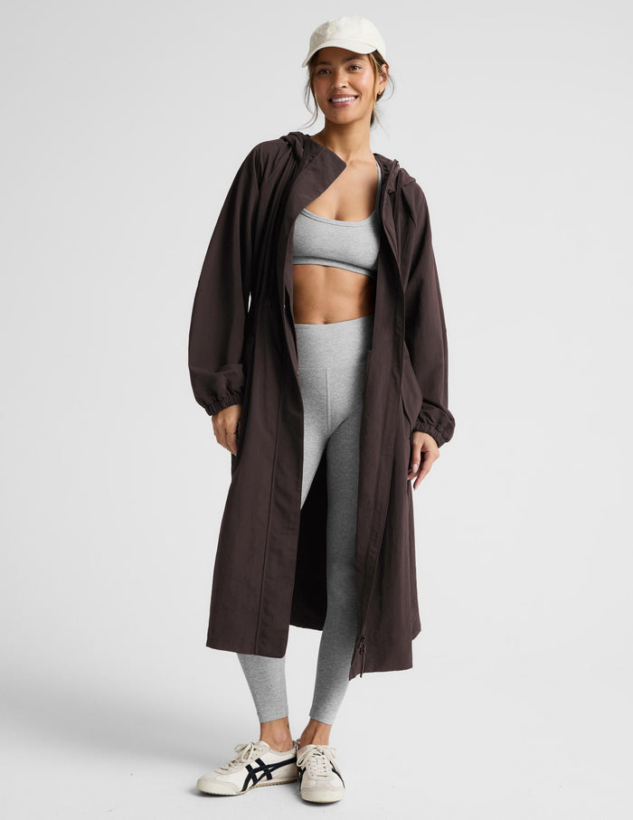 A woman modeling a dark brown hooded longline coat with drawstrings and large front patch pockets, paired with light gray leggings and white sneakers, against a light gray studio background.