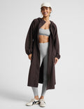 A woman modeling a dark brown hooded longline coat with drawstrings and large front patch pockets, paired with light gray leggings and white sneakers, against a light gray studio background.