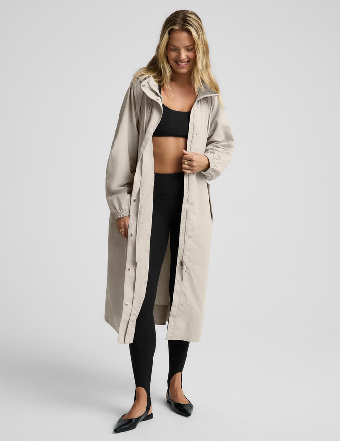 A woman modeling a long, light beige hooded raincoat over a black crop top, black leggings, and black flats, photographed against a solid light gray studio background.