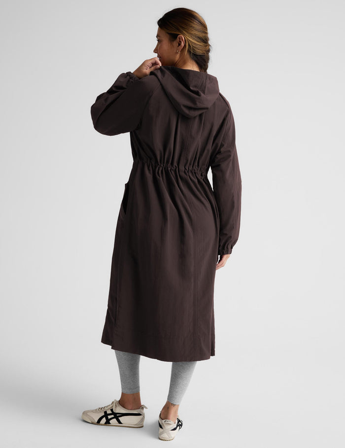 A woman modeling a dark brown hooded longline coat with drawstrings and large front patch pockets, paired with light gray leggings and white sneakers, against a light gray studio background.