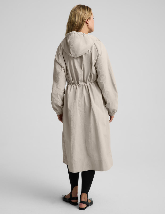 A woman viewed from the back wearing a light beige hooded raincoat with a drawstring waist, black leggings, and black sandals against a light gray studio background.