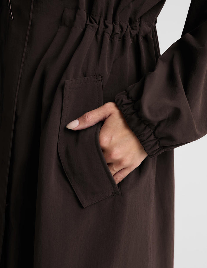A woman modeling a dark brown hooded longline coat with drawstrings and large front patch pockets, paired with light gray leggings and white sneakers, against a light gray studio background.
