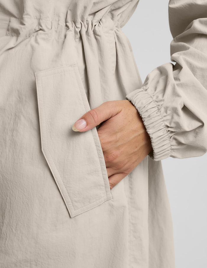 Close-up of a light beige or oatmeal-colored garment with an elasticized waistband and a side pocket, shown on a person with one hand inserted into the pocket against a plain light gray background.