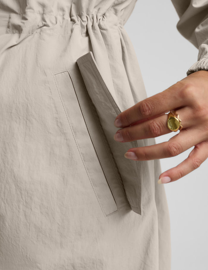 Close-up of a person wearing a light beige or oatmeal-colored garment with a gathered waistband and a side pocket, with a hand resting near the pocket. The person is also wearing a gold ring with a green stone.