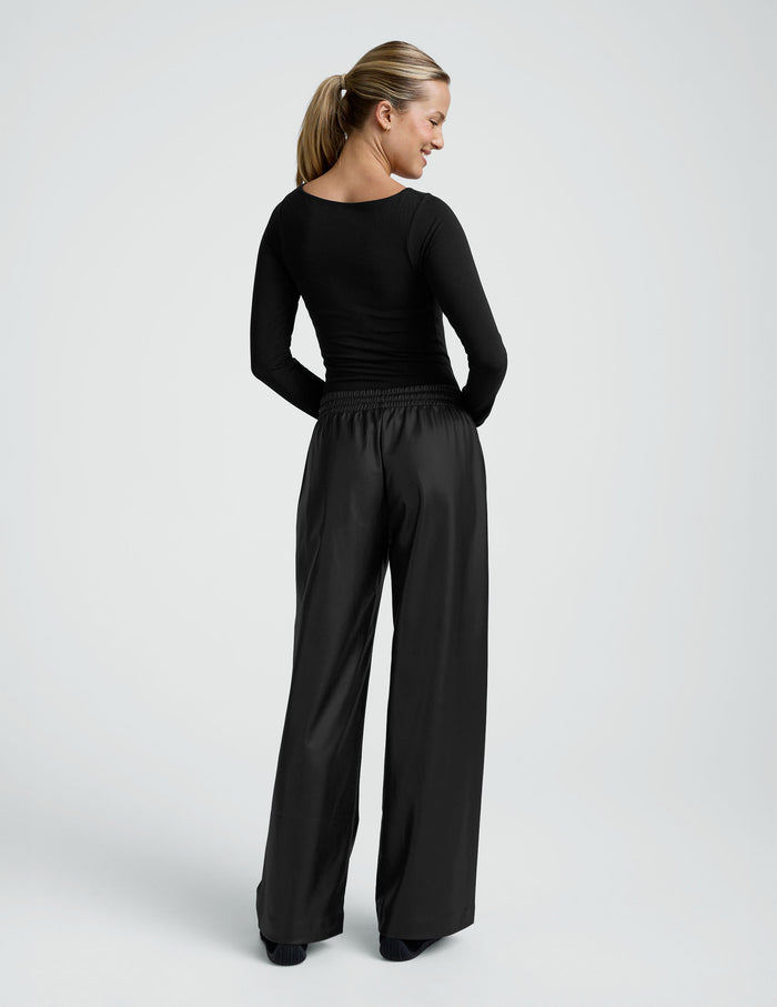 Rear view of a model wearing black wide-leg Faux Leather Pant with a gathered elastic waistband and sleek, full-length fit.