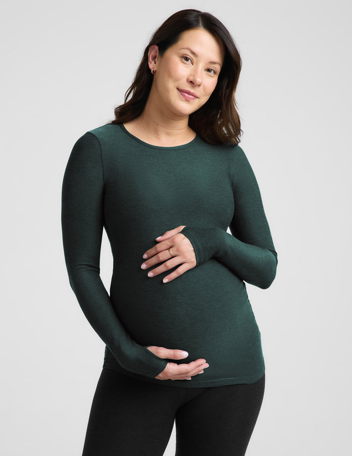 A pregnant woman wearing a dark forest green long-sleeve maternity top and black leggings, standing against a light gray studio background.