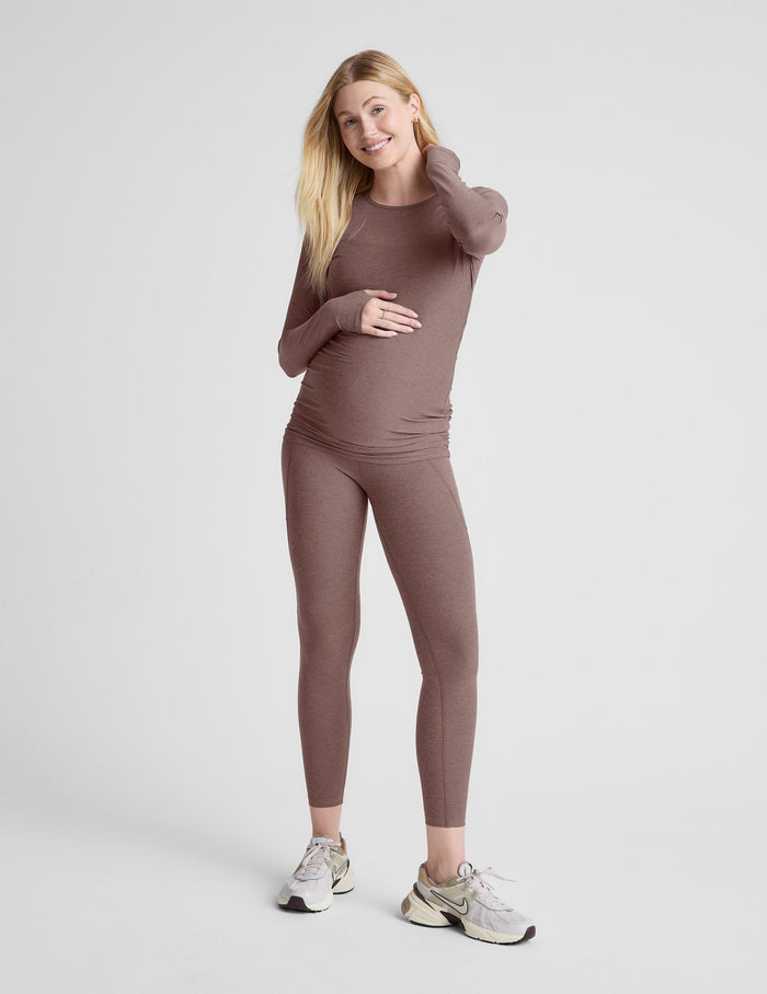 Woman wearing a brown maternity long-sleeve top and leggings against a light gray background.