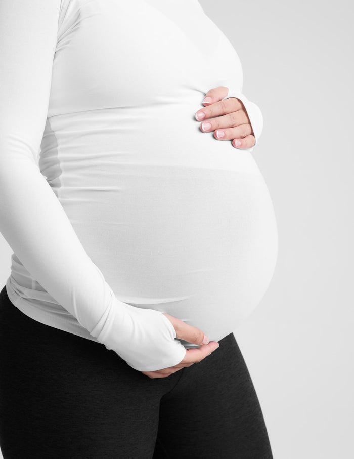 A pregnant woman wearing a white long-sleeve maternity top and black leggings, standing against a light gray studio background.