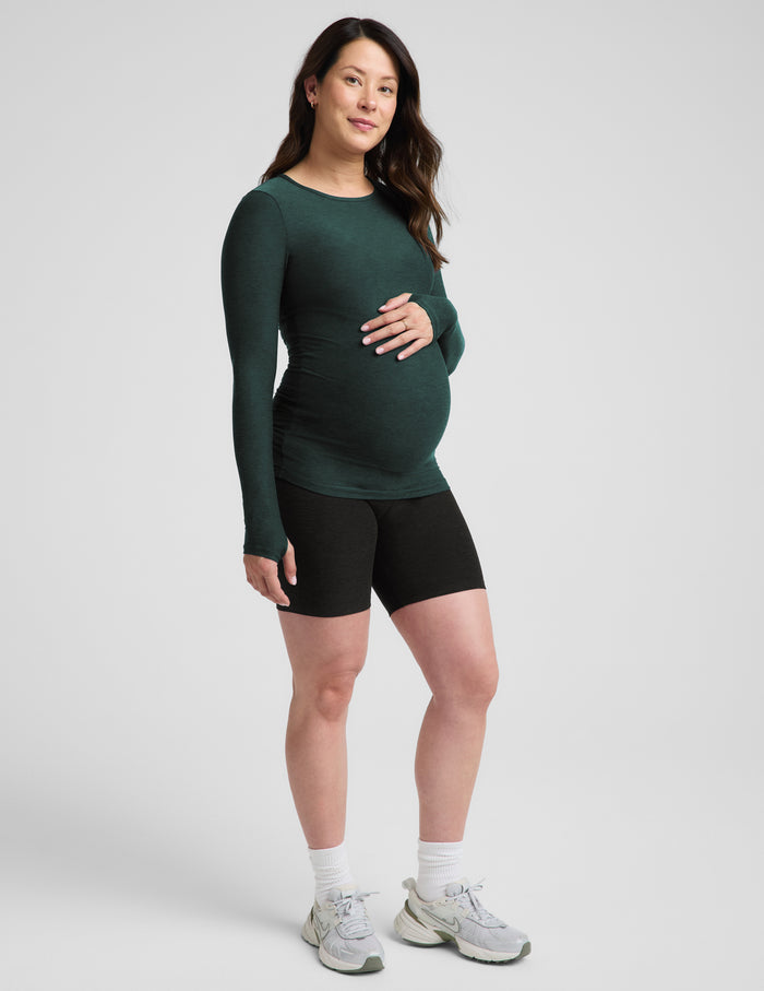 A pregnant woman modeling a dark green long-sleeve maternity top and black athletic shorts against a light gray studio background.