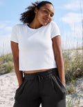 A woman wearing a white short-sleeve cropped t-shirt and black drawstring sweatpants, standing outdoors on a sandy beach with dune grass and a blue sky in the background.