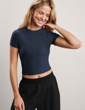 A woman wearing a navy blue short-sleeve crop top and black pants, posing against a light gray studio background.
