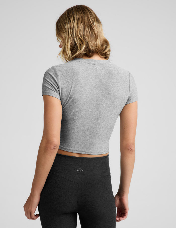 A woman wearing a light heather gray short-sleeve crop top and black bottoms, photographed against a plain light gray studio background.